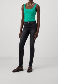 Grön åtsittande linne, svarta skinny jeans med lätt slitage och platta sandaler. Modellen står och visar upp outfiten mot en neutral bakgrund.