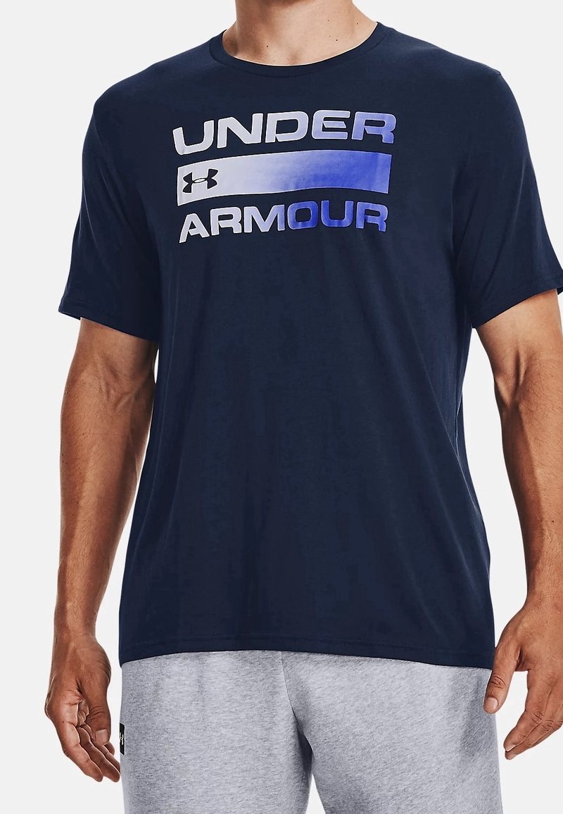Námořnická modrá tričko s krátkým rukávem, na kterém je logo "UNDER ARMOUR" v gradientním písmu. Vyrobeno z měkké látky s volným střihem, ideální k šedým šortkám.