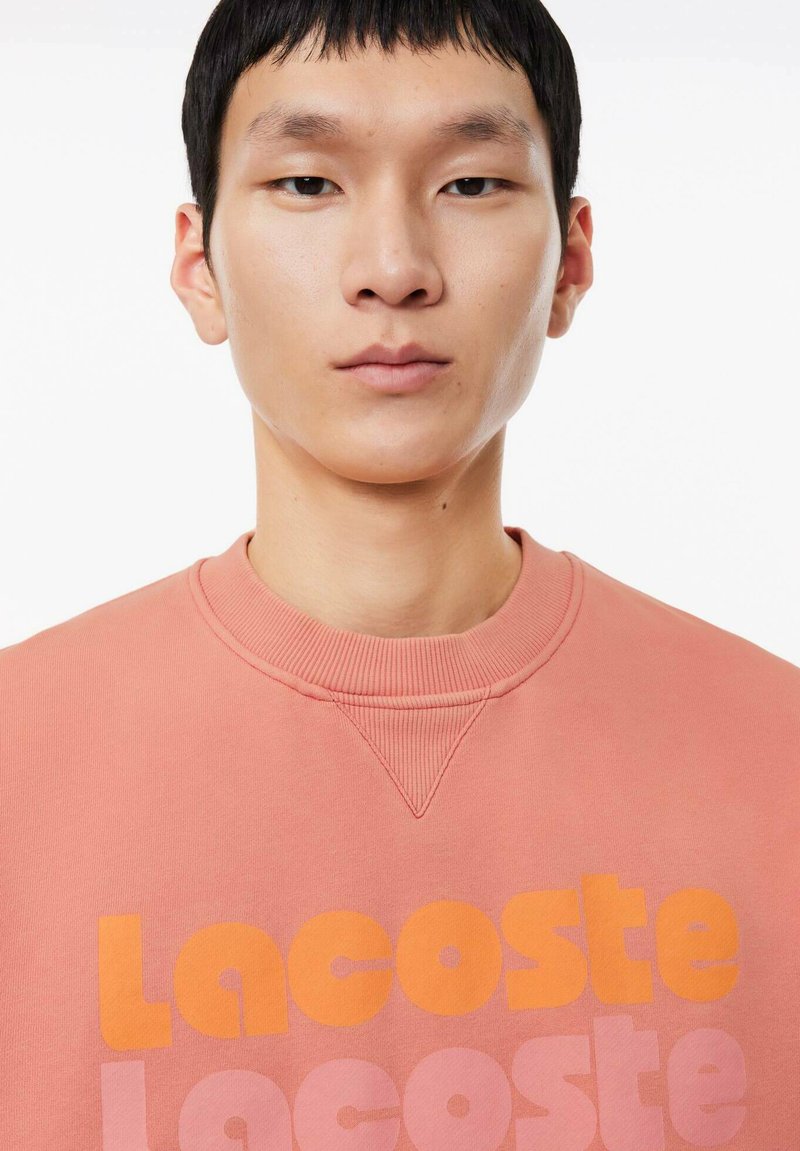 Felpa rosa chiaro con colletto a coste, caratterizzata da un audace testo arancione "Lacoste" sul davanti e una texture di cotone liscia.