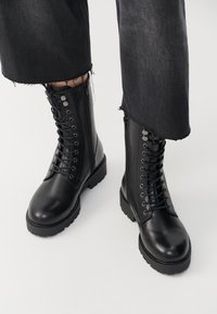 Zwarte enkelboots met een zachte leren afwerking, vetersluiting aan de voorkant en een zijrits. Chunky zool en gedetailleerde veterogen voegen textuur toe.