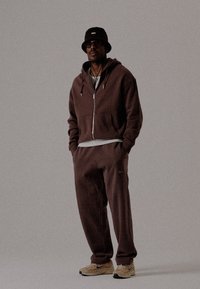 Homme portant un hoodie marron foncé et un pantalon de survêtement, des baskets beiges, un chapeau seau noir et des lunettes, se tenant les mains dans les poches devant un fond uni.