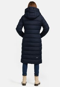 Navy puffer coat with a hood, featuring vertical seams and a logo detail, extending to mid-calf, paired with blue jeans and beige boots.