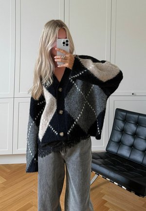 Kootud ülemahutav kardigan musta, halli ja beeži rombikujulise mustriga, millel on suured nööbid ja tekstuuriga ääreosa, kombineeritud hallide laiade pükstega.