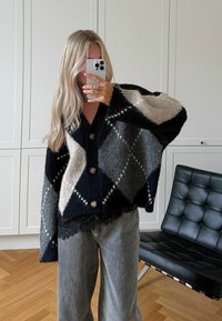 Dzianinowy, oversizeowy kardigan w czarno-szaro-beżowy wzór w romby, z dużymi guzikami i fakturowanym dołem, zestawiony z szarymi szerokimi spodniami.