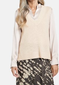 Donna che indossa un gilet di maglia beige sopra una camicia di raso color crema e una gonna con stampa animale nera e beige, in piedi contro uno sfondo bianco.