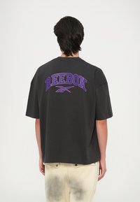 Camiseta negra de gran tamaño con texto en morado "REEBOK" en la parte posterior, mangas cortas y un escote redondo. El modelo lleva pantalones crema con acentos oscuros.