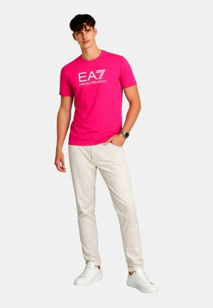 Uomo che indossa una t-shirt EA7 Emporio Armani rosa acceso, pantaloni beige chiaro, sneakers bianche e un orologio da polso, in piedi davanti a uno sfondo semplice.