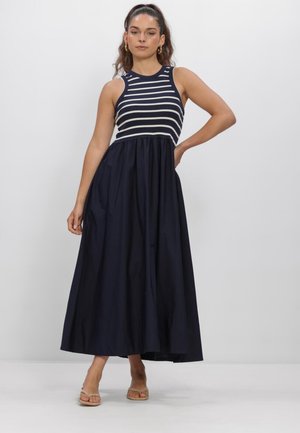 Frau in einem dunkelblauen ärmellosen Kleid mit weißen horizontalen Streifen auf der oberen Hälfte und einem einfarbigen, fließenden Rock, stehend mit einer Hand in der Hüfte.