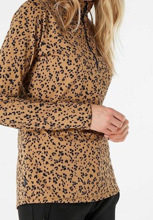 Vrouw draagt een bruin topje met lange mouwen, zwart luipaardprint en een halve rits, handen in elkaar gevouwen ter hoogte van de taille, tegen een witte achtergrond.