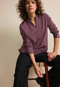 Løs-fit mauve bluse med V-hals og lange ærmer, parret med sorte bukser med striber. Modellen sidder på en træstol.
