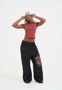 Croptop in roestkleur met zilveren floral ontwerp, gecombineerd met losse zwarte denim broek met geborduurde rozen en accentpatches.