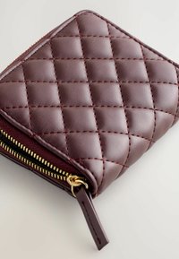 Portefeuille en cuir bordeaux avec texture matelassée. Doté d'une fermeture éclair dorée et de coutures rouges. Forme carrée compacte avec un profil mince.