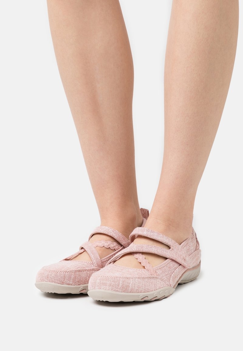 Chaussures style Mary Jane rose clair avec brides velcro et tissu texturé portées pieds nus sur un fond uni.