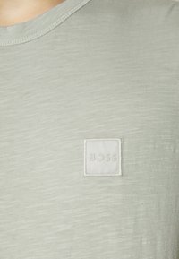 Ljusgrön T-shirt i mjukt tyg, med en fyrkantig vit etikett med "BOSS" broderad, rund halsringning och subtil textur.