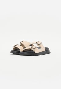 Beige suedinesandaler med to justerbare stropper og sølvspenne, med en svart gummisåle og et minimalistisk design.