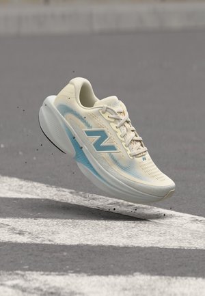 Chaussure de course New Balance beige et bleue en l'air au-dessus de l'asphalte avec des bandes blanches et de petits débris autour.