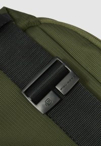 Borsa in tessuto verde con superficie strutturata, dotata di tracolla nera regolabile e fibbia metallica con dettagli di branding.