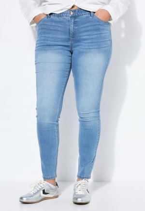 Person iført lyseblå skinny jeans med hænderne i lommerne og sølv- og hvide sneakers, stående mod en hvid baggrund.