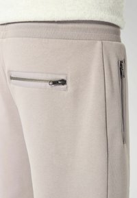 Jasnoszare dresy z ribbowanym paskiem w talii, z zamykaną na zamek kieszenią boczną oraz gładką teksturą. Prosty i funkcjonalny design.