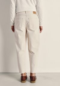 Persona vistiendo jeans rectos de color blanco hueso, zapatos de cuero marrón y una blusa de manga larga color crema, de pie frente a un fondo liso.