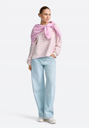 Ženska v roza puloverju z rumenimi črtami in roza majico, zavezano okoli ramen, svetlo modre široke kavbojke in bele superge.