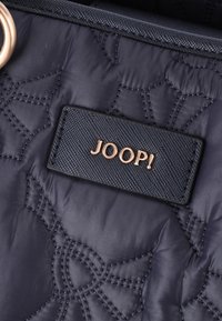 Laavanssin väritetty tikattu kankainen laukku, jossa on teksturoitu muotoilu, musta tagi, jossa on kultakirjoitus "JOOP!" ja pyöreät kiinnityselementit.