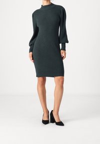Abito a maglia a coste di colore verde scuro con un collo alto, maniche lunghe a sbuffo e una silhouette aderente, abbinato a scarpe con tacco a blocco nere.