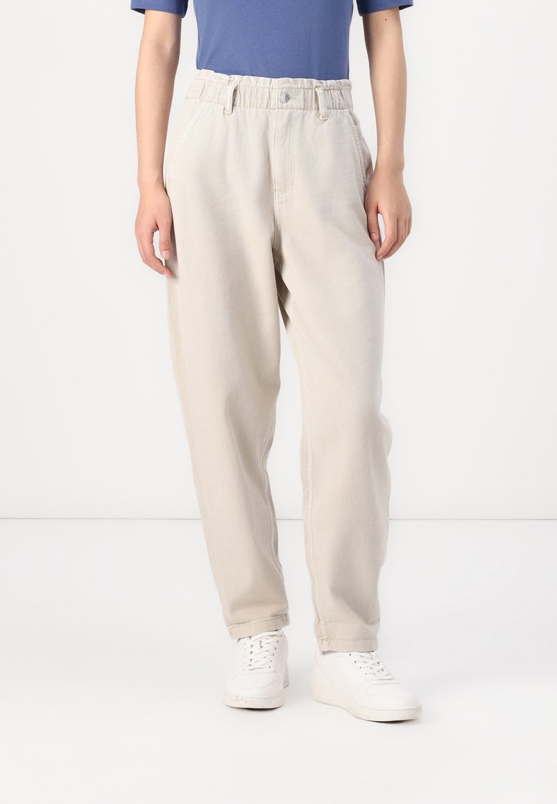 Pantalones de algodón color beige claro con cintura elástica, bolsillos laterales y un corte relajado, combinados con zapatillas blancas.