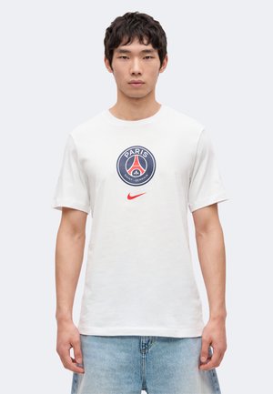 Homme portant un t-shirt blanc Paris Saint-Germain avec un logo bleu marine et rouge et un swoosh Nike rouge, associé à un jean bleu clair.