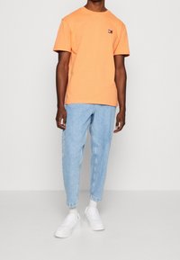 Oranžové bavlněné tričko s kulatým výstřihem a malou nášivkou. V kombinaci se světle modrými džíny a bílými teniskami vytváří ležérní vzhled.