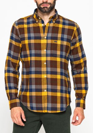 Uomo che indossa una camicia a quadri gialla, marrone, blu e grigia con maniche lunghe e pantaloni verde scuro.