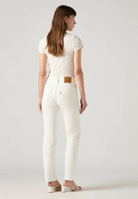 Witte denim jeans met een hoge taille, achterzakstiksels en een leren patch in bruin. Het model draagt een lichtgepatroneerd topje en beige hakken.