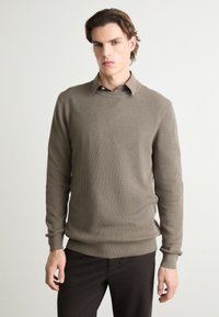 Suéter de punto acanalado beige con escote redondo y mangas largas, combinado con pantalones oscuros. La textura de la tela es suave y sutil.