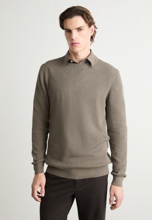 Pull en tricot côtelé beige avec un col rond et des manches longues, associé à un pantalon sombre. La texture du tissu est douce et subtile.