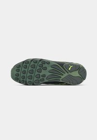 La semelle de la chaussure de sport noire présente un motif de grip en caoutchouc texturé avec des accents verts, des lignes courbes et un logo bien en vue.