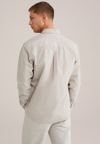 Gestreepte beige shirt met lange mouwen, achterkant met plooi detail en knoopmanchetten; gedragen met lichtgekleurde broeken. Zachte textuur, casual ontwerp.