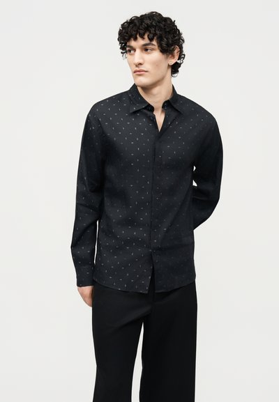 Νέος άνδρας με κατσαρά μαλλιά που φοράει μαύρο πουκάμισο με κουμπιά με λεπτό λευκό μοτίβο και μαύρο παντελόνι, στέκεται μπροστά σε απλό φόντο.