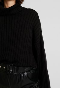 Pull à col roulé noir à côtes avec manches longues. Texture douce, coupe loose. Porté avec un pantalon en cuir noir taille haute et une large ceinture.