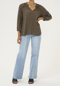 Blouse marron à pois avec un col en V et un détail plissé, associée à un jean évasé bleu clair et des talons ouverts noirs.