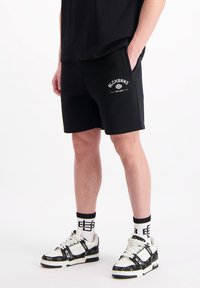 Zwarte katoenen shorts met zijzakken, voorzien van witte bedrukte tekst. Gepaard met witte en zwarte sneakers en gestreepte crew-sokken.