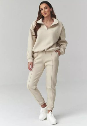 Kobieta z długimi włosami, ubrana w beżową bluzę z półzamkiem i dopasowane spodnie dresowe, stojąca z jedną ręką w kieszeni, ubrana w białe trampki.