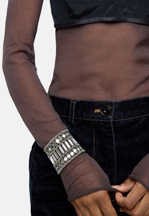 Brązowa, przezroczysta bluzka z długim rękawem nałożona na czarną, krótką bluzkę, w połączeniu z ciemnoniebieskimi dżinsami. Wielowarstwowa srebrna bransoletka z koralikami i akcentami.