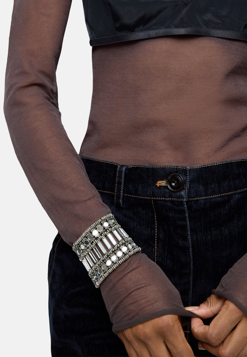 Brązowa, przezroczysta bluzka z długim rękawem nałożona na czarną, krótką bluzkę, w połączeniu z ciemnoniebieskimi dżinsami. Wielowarstwowa srebrna bransoletka z koralikami i akcentami.