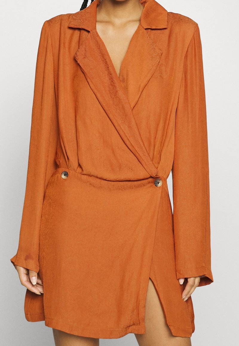 Femme portant une robe portefeuille orange rouille à manches longues, col tailleur, et détails de boutons à la taille, avec une fente haute sur la cuisse.