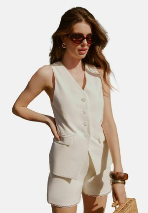 Off-white mouwloos vest met vijf knopen, zijzakken; bijpassende shorts met een decoratieve zoom. Accessoires zijn onder andere schildpadbril en armbanden.