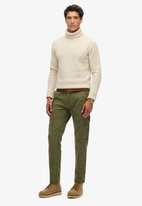 Beige coltrui, olijfgroene cargobroek met zakken, bruine riem, beige veterschoenen, staande figuur met een informele houding.