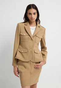 Mujer vistiendo una chaqueta beige a medida con cuatro bolsillos abotonados y una falda lápiz a juego, de pie frente a un fondo claro y liso.