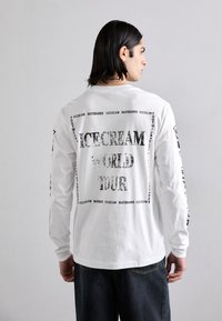 White long-sleeve shirt featuring black text graphics, including "ICECREAM WORLD TOUR," with contrasting textured print and a relaxed fit.