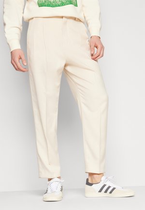 Man wearing cream-colored tailored pants with crease lines, paired with white low-top sneakers with black stripes.