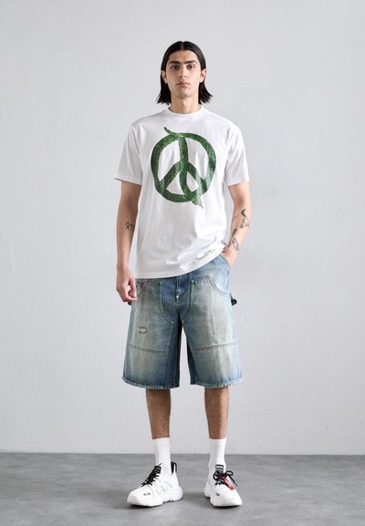 Camiseta de algodón blanca con un símbolo de paz estampado en patrón de serpiente verde, combinada con unos pantalones cortos de mezclilla azul holgados y zapatillas blancas.